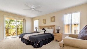 A bedroom with a large bed, a ceiling fan, a nightstand with a lamp, a sofa, and a balcony with a railing.