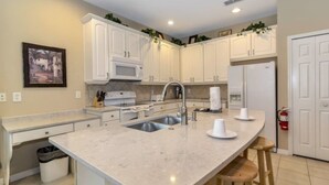 A modern kitchen with white cabinetry, a large island with stools, stainless steel appliances, and a tiled backsplash. - Watersong - Luxurious 5bd/4.5ba Pool Home #5wr510 (Davenport)