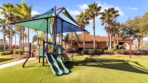 A playground with a slide and climbing structures, surrounded by palm trees and a building with a red-tiled roof.