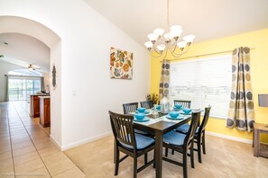 Uma sala de jantar com uma mesa posta para seis pessoas, seis cadeiras, um candelabro, uma janela com persianas e uma parede amarela.