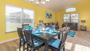 A dining area with a table set for six, surrounded by chairs with blue cushions. There is a chandelier above the table, and the walls are painted yellow.