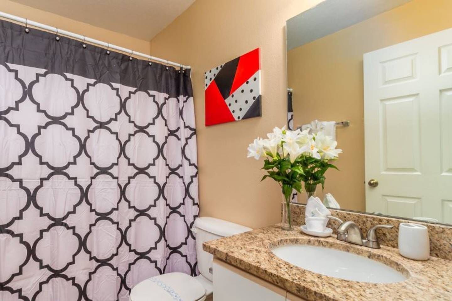 A bathroom with a patterned shower curtain, a sink with a granite countertop, a vase of white flowers, and a mirror reflecting a door.