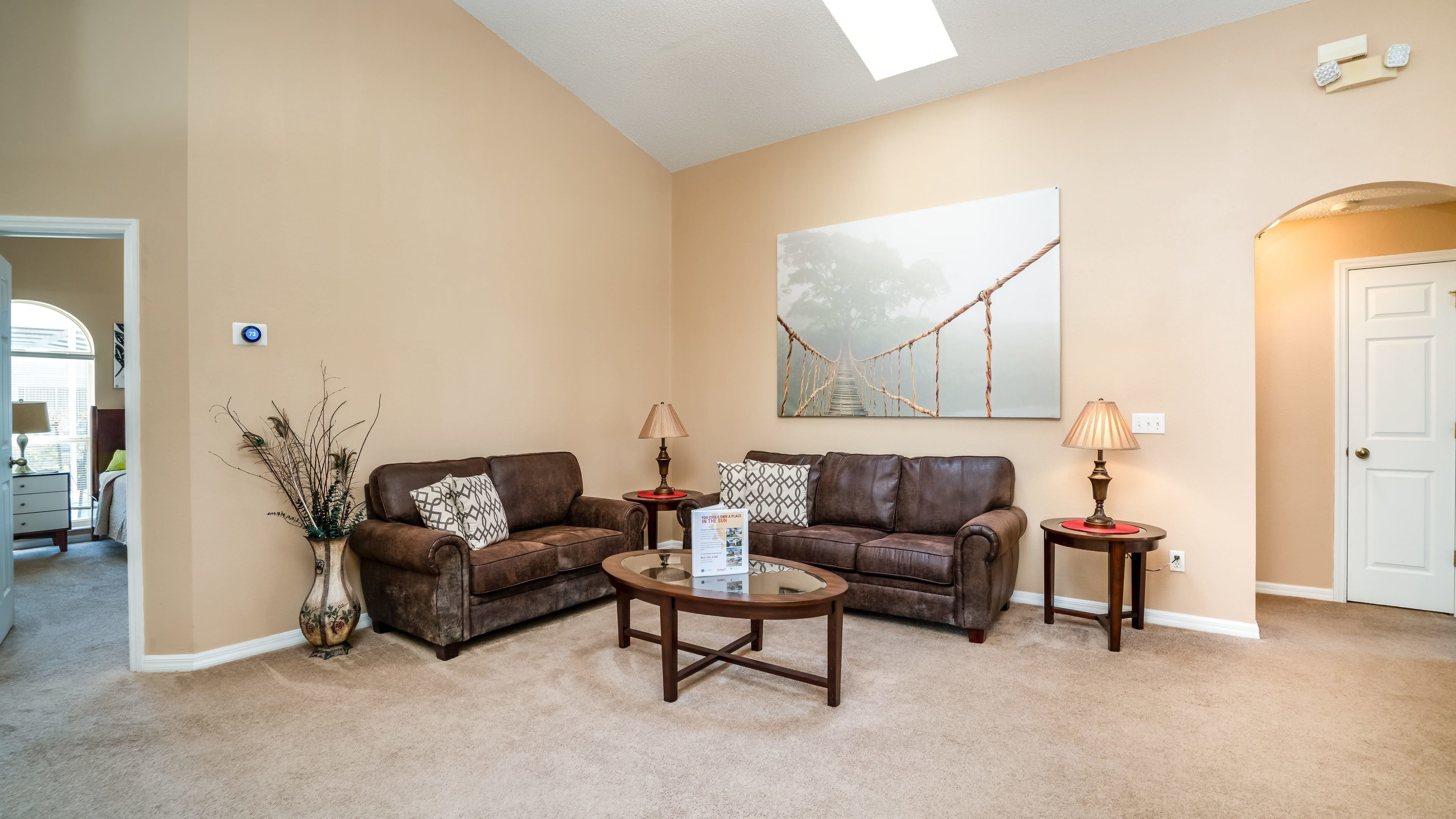 A living room with two brown leather sofas, a glass coffee table, and two side tables with lamps. A large painting of a bridge is on the wall, and a door is visible on the right.