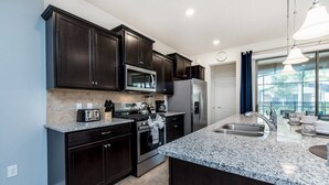 A modern kitchen with dark cabinetry, granite countertops, stainless steel appliances, and a large island with a sink. - Solterra Resort - Brand NEW 5bed Pool Home #5st417 (Davenport)