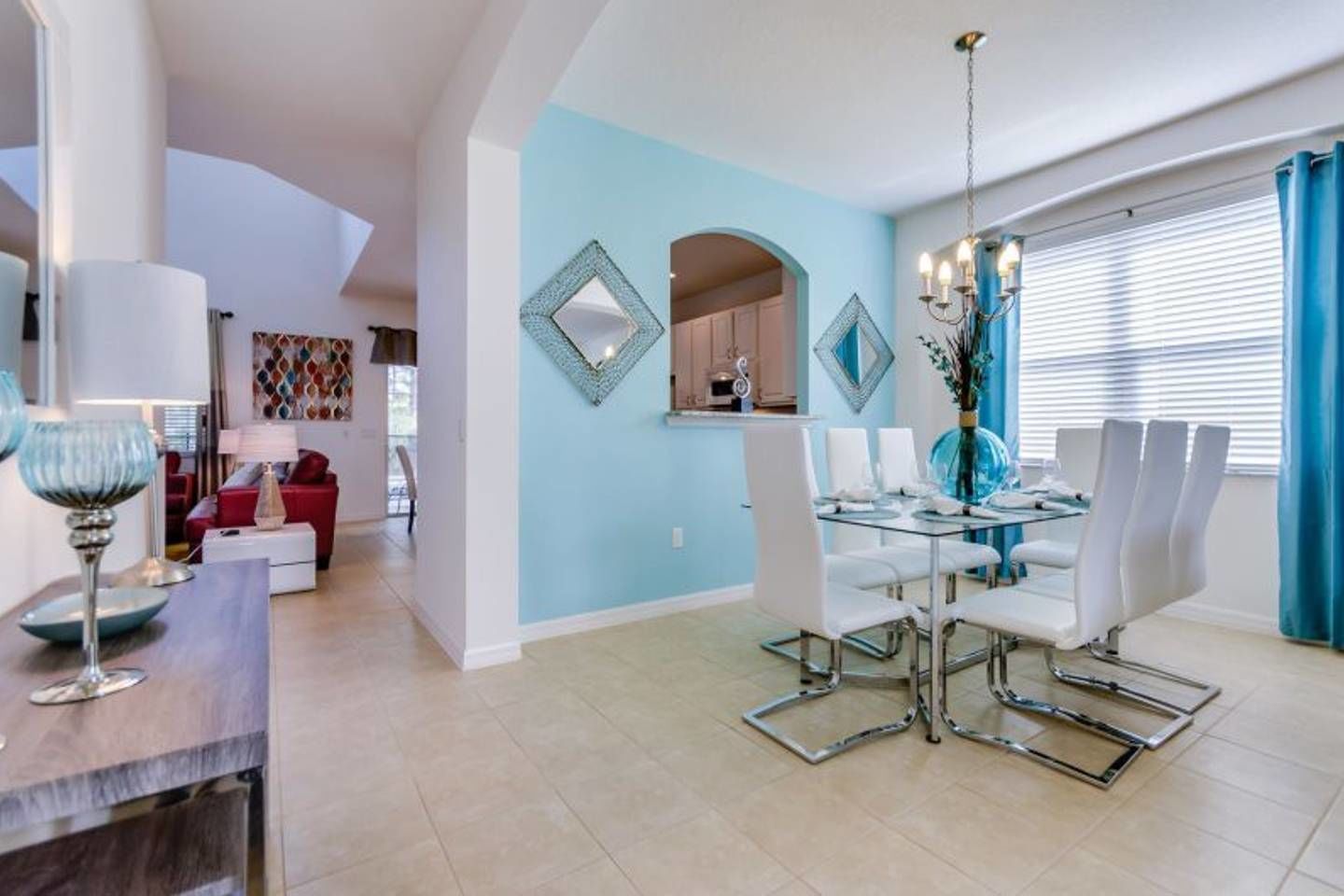 A modern dining area with a glass table and white chairs, a kitchen in the background, and a blue accent wall.