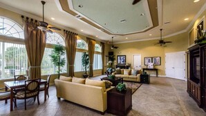 A spacious living room with a large sofa, a dining table with chairs, and a coffee table. There are large windows with curtains, a ceiling fan, and a television on the wall. - Watersong - 6bd/5.5ba Pool Home #6wr120 (Davenport)