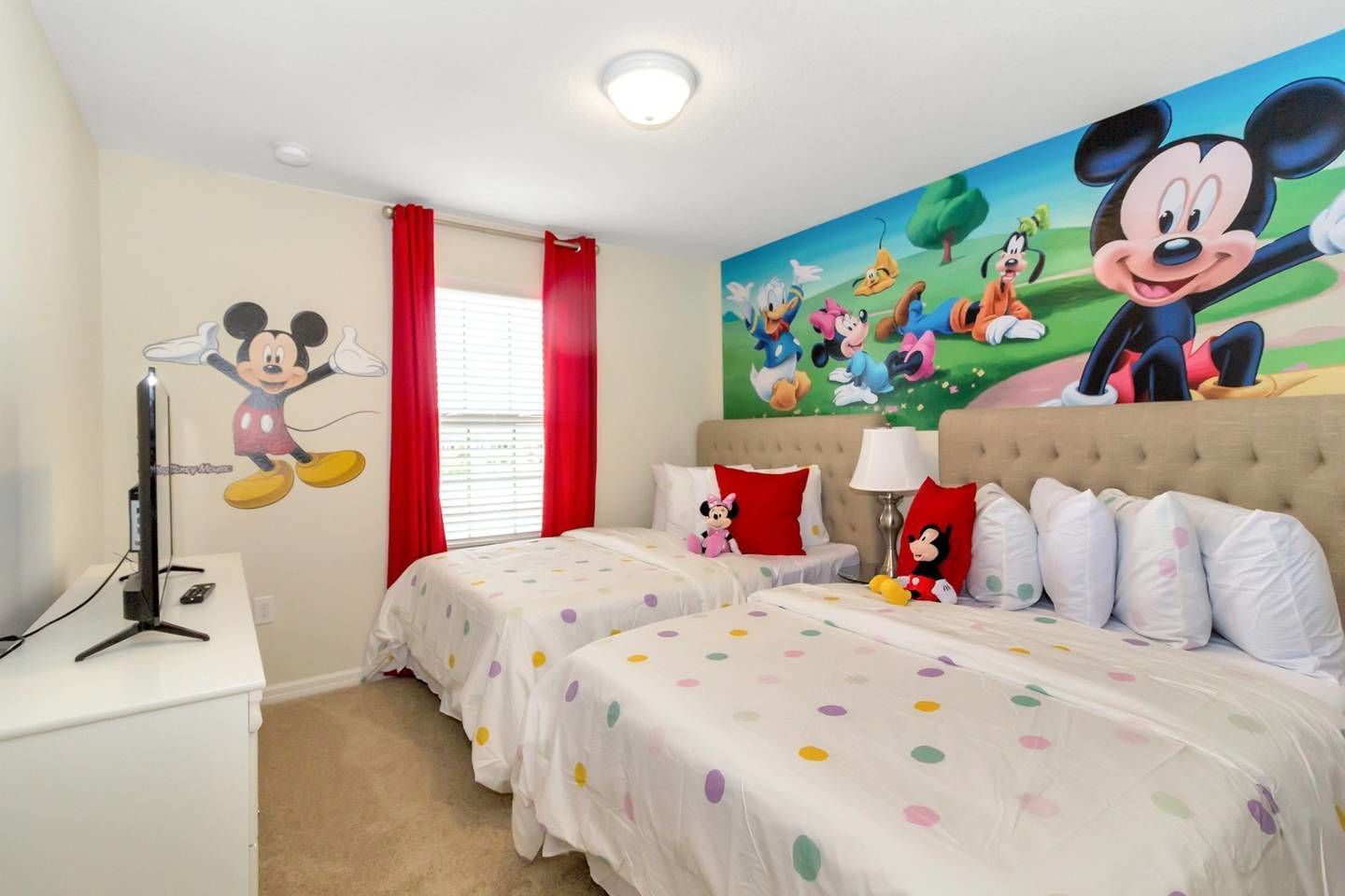 Habitación de hotel con dos camas, un gran mural con temática de Disney, cortinas rojas y un televisor sobre un soporte blanco.