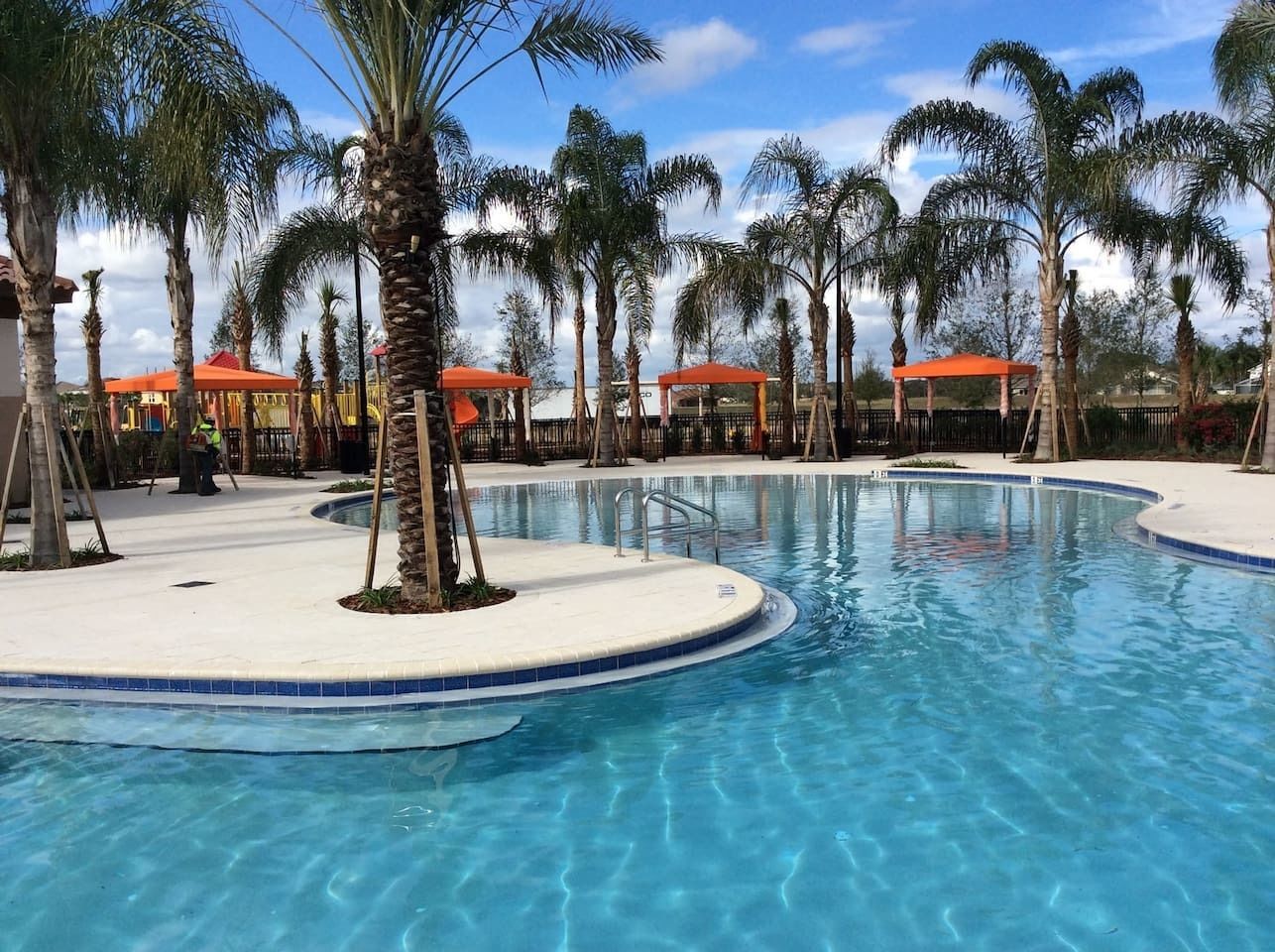 A swimming pool with palm trees and red-roofed structures in the background.