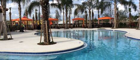 A swimming pool with palm trees and red-roofed structures in the background.