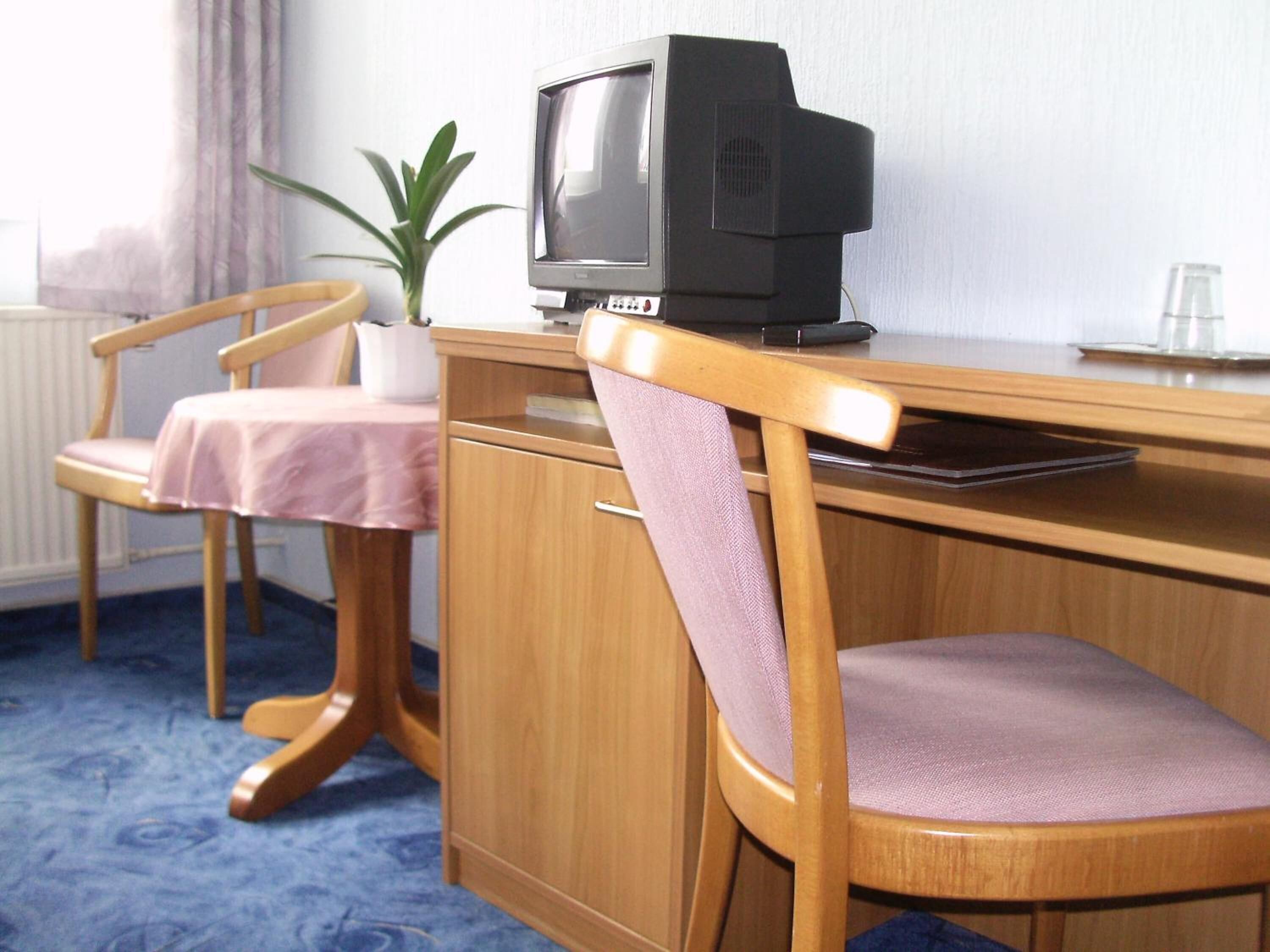 Una habitación con un escritorio de madera, televisión, una silla, una mesita con mantel rosa y una planta en maceta.