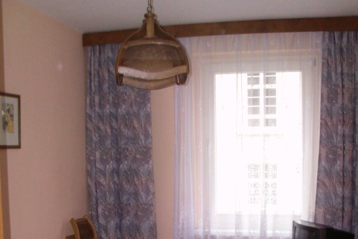A single bed with a wooden headboard, a bedside table, a chair, a television, and a window with curtains.