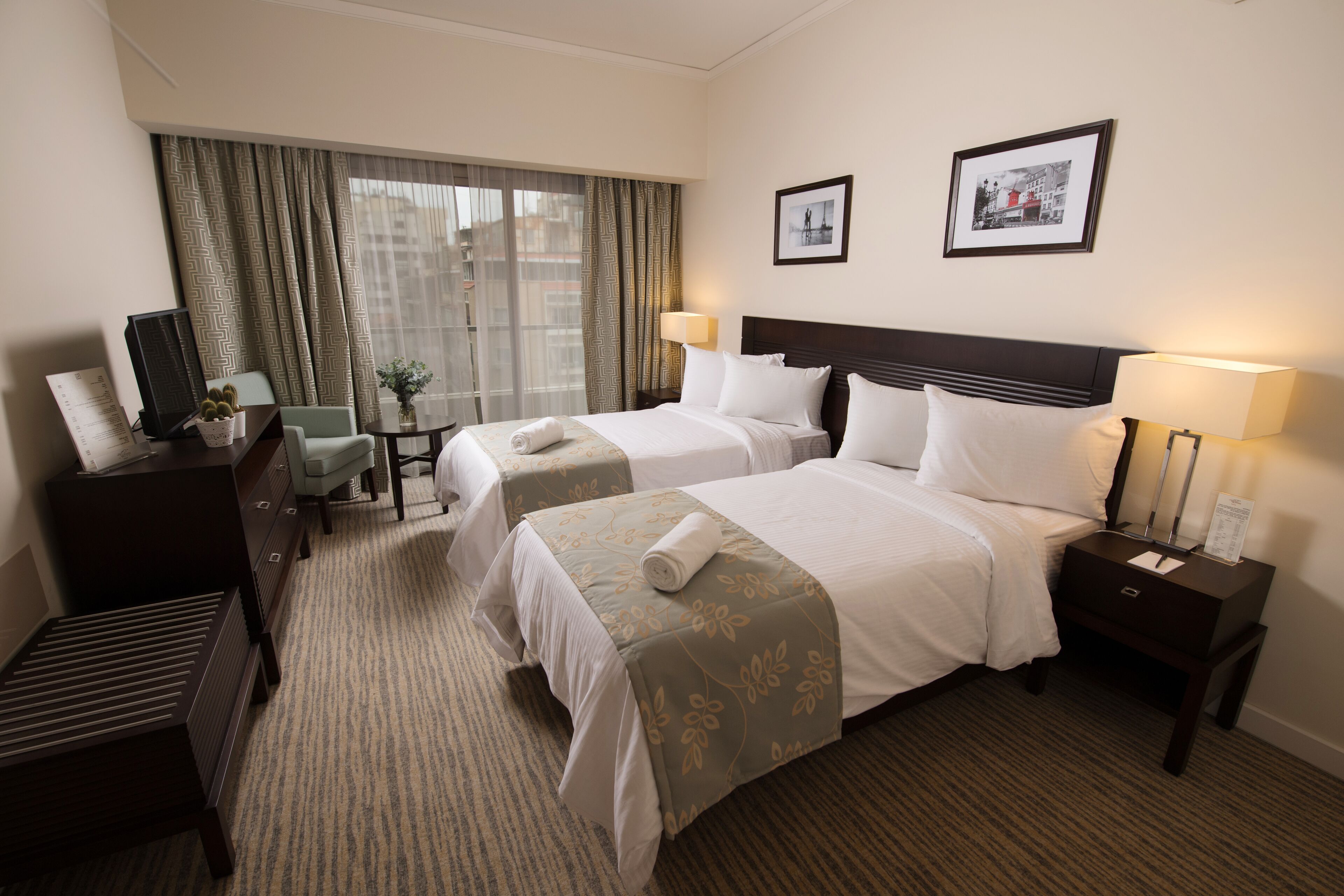 A hotel room with two beds, a desk, a chair, a small table with a plant, and a window with curtains.