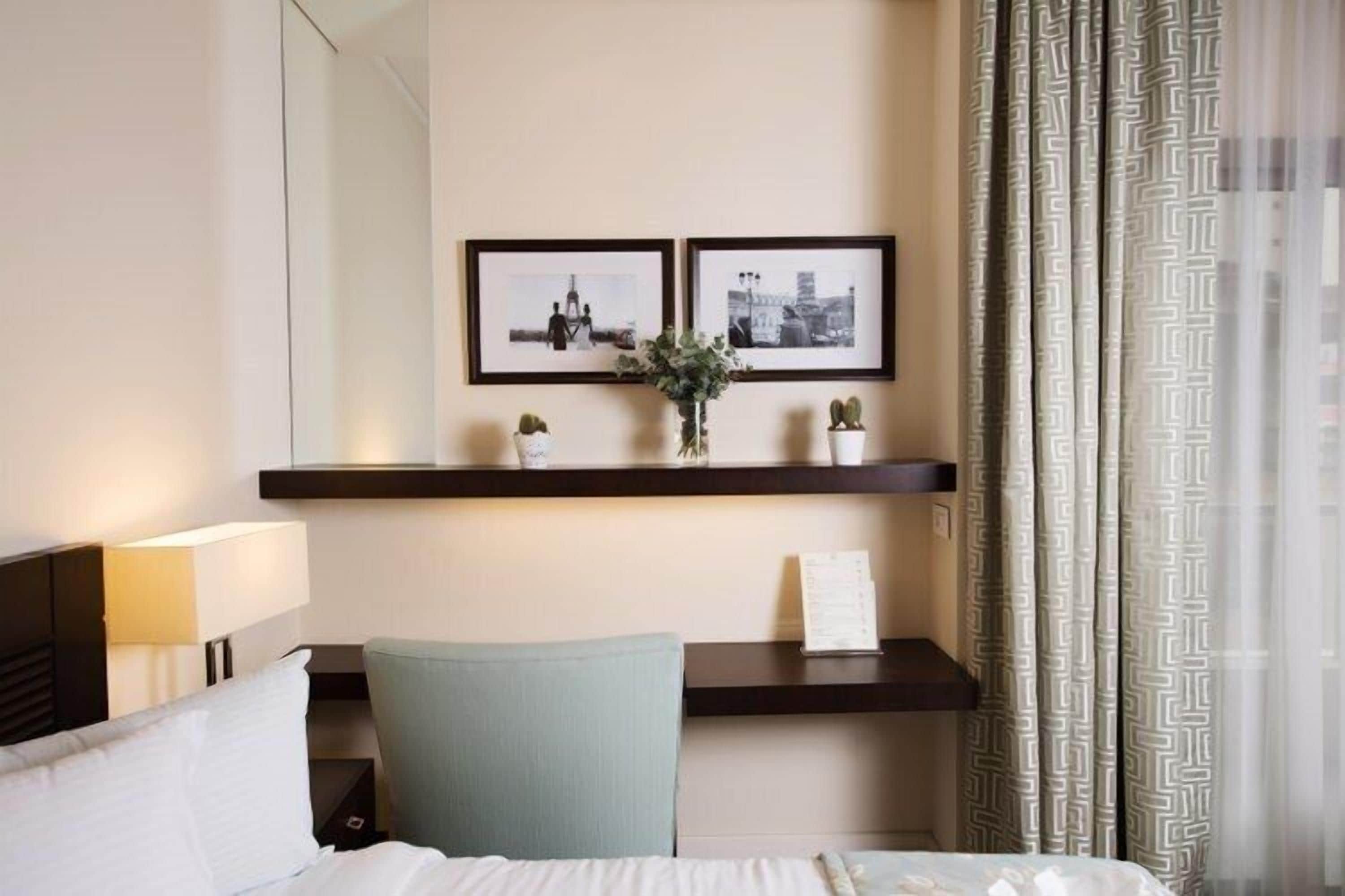 Una habitación de hotel con una cama, un escritorio con lámpara, dos cuadros en la pared y una ventana con cortinas estampadas.