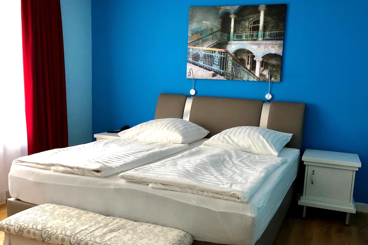A neatly made double bed with white linens, a bench at the foot of the bed, a bedside table, and a wall-mounted picture of a bridge.