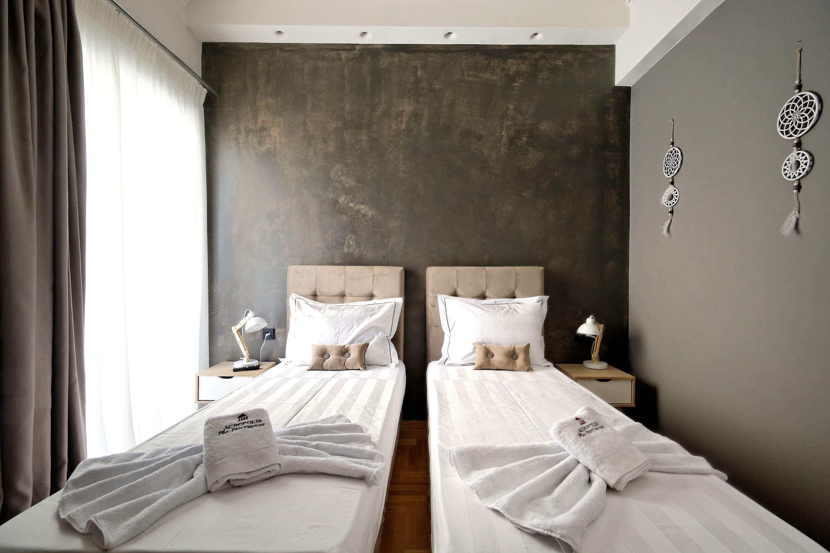 A hotel room with two beds, each with a white bedspread and pillows, a bedside table with a lamp, and a large window with curtains.
