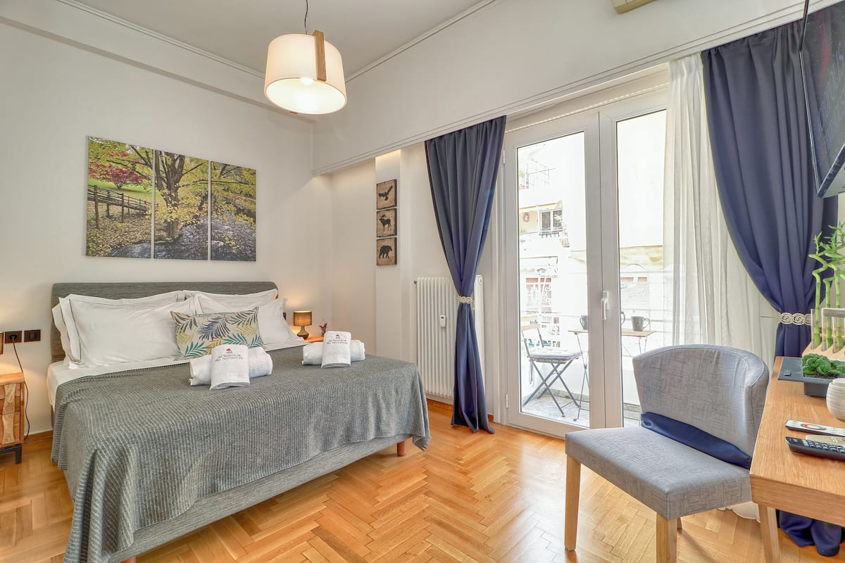 A modern hotel room with a bed, a chair, a table, and a balcony with curtains.