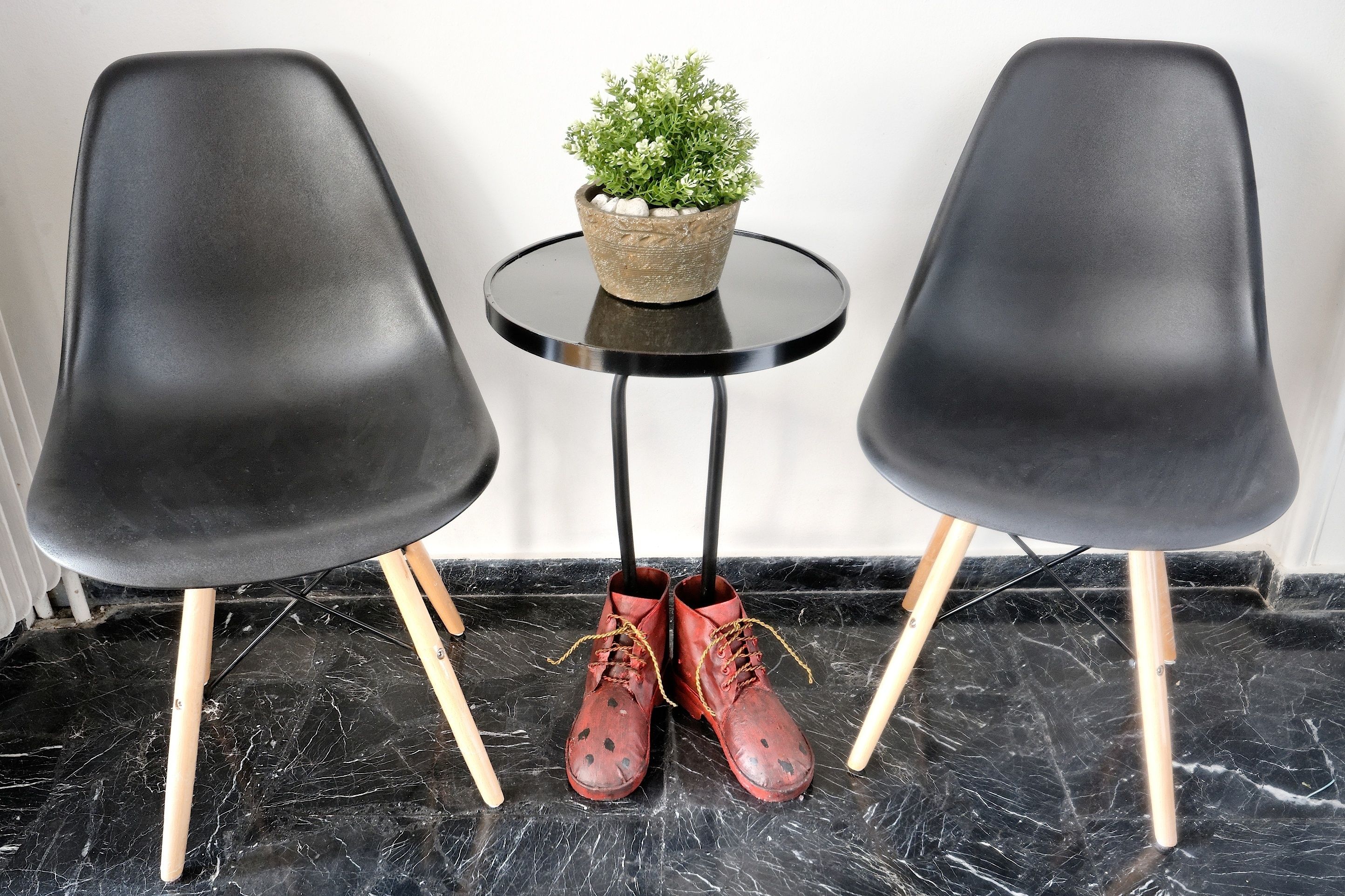 Deux chaises noires aux pieds en bois, une petite table ronde ornée d’un pot de fleurs et une paire de chaussures rouges à lacets, posées sur le sol.