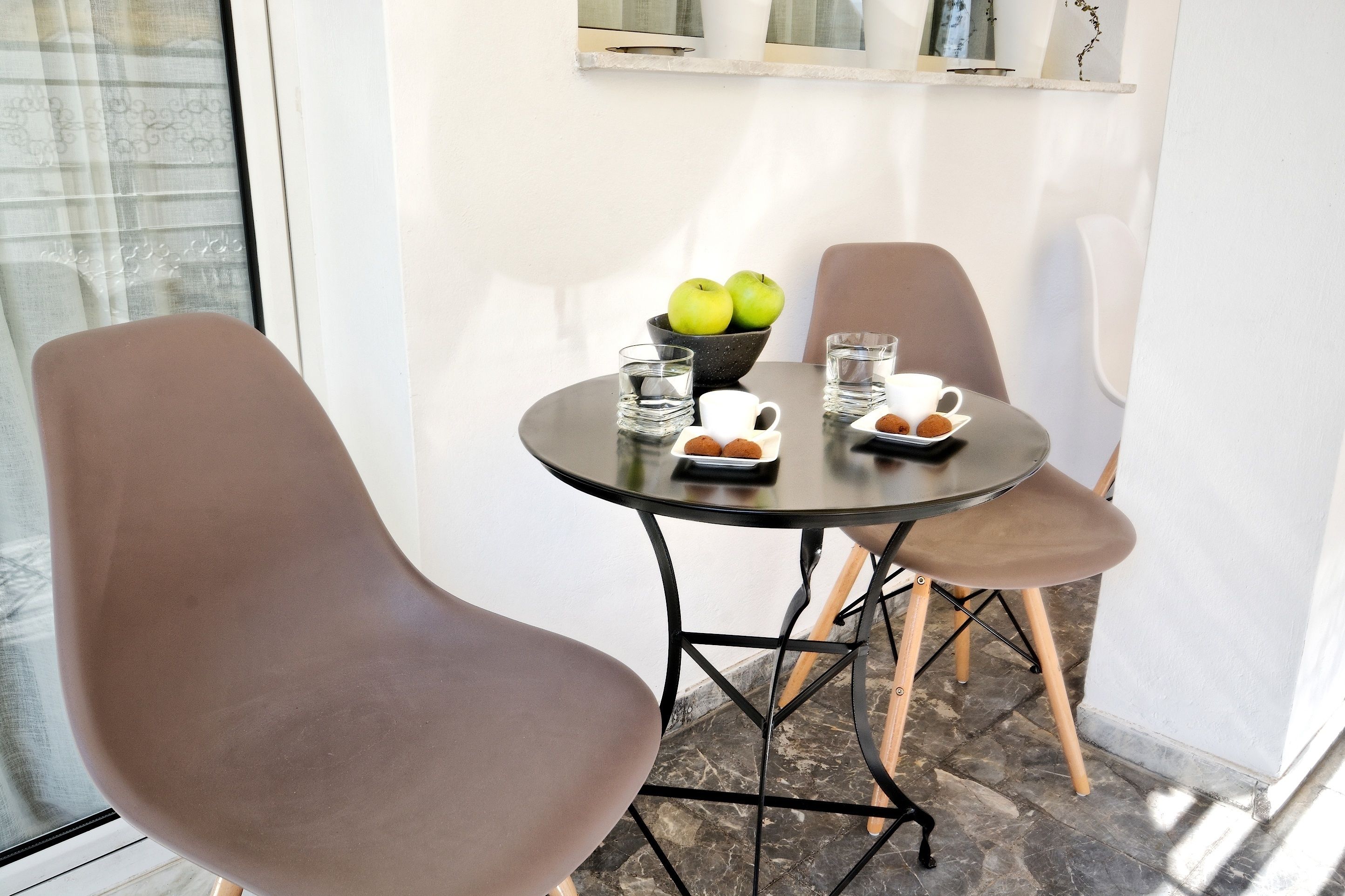 A small dining area with a round table, two chairs, a bowl of green apples, two glasses, a white mug, and a plate with cookies.