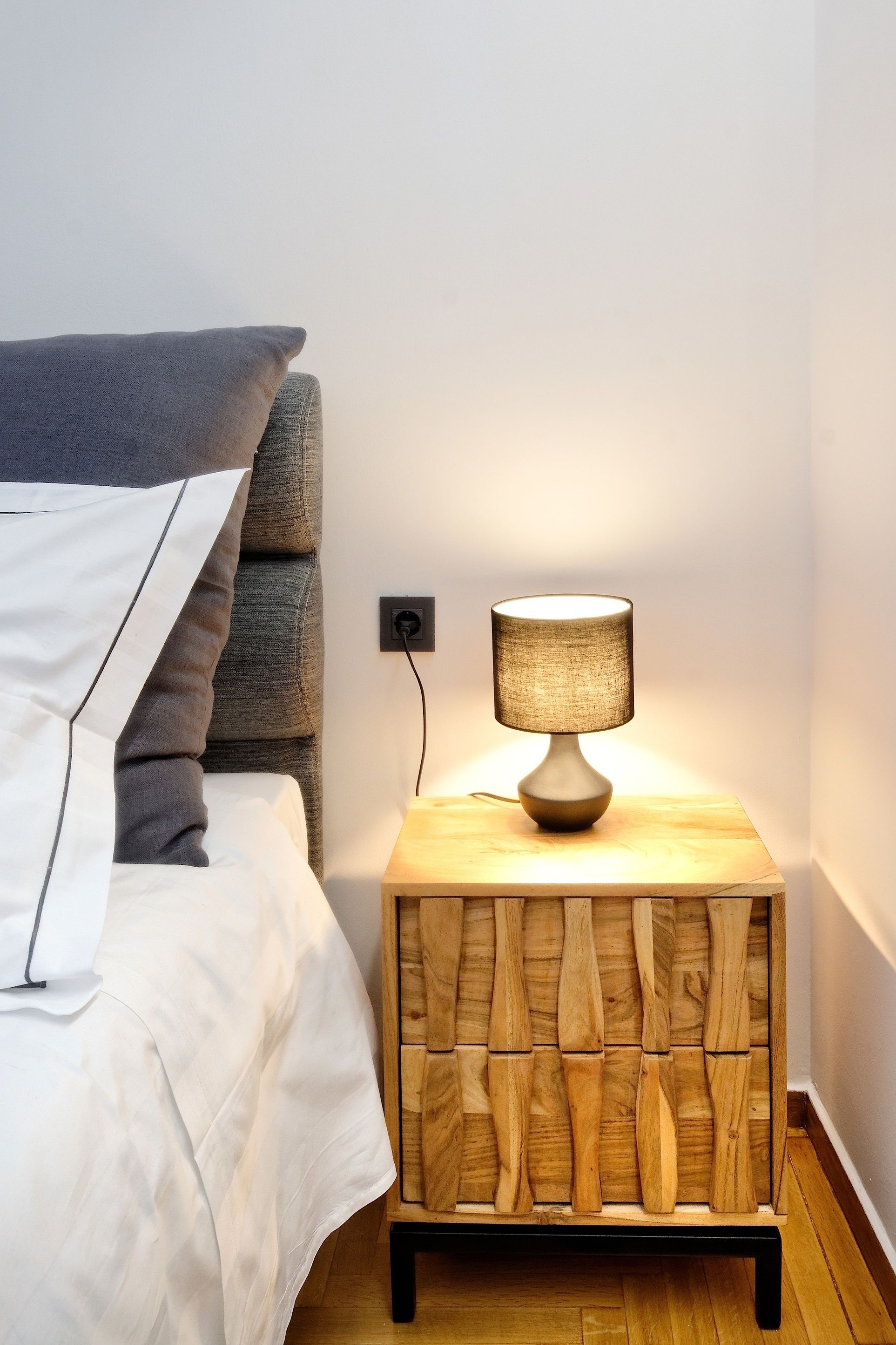 A neatly made bed with a gray and white pillow, a wooden nightstand with a lamp, and a wall socket.