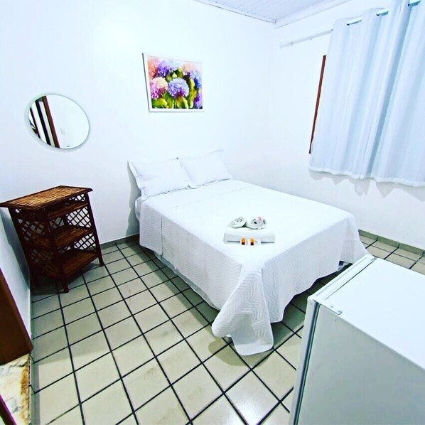 A small, clean room with a bed, a small table, a mirror, and a painting on the wall. - Pousada Oliveira (Cairu)