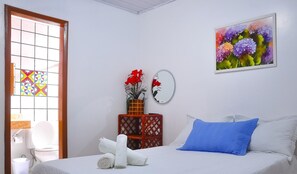A hotel room with a bed, a small table with a vase of red flowers, a painting of flowers on the wall, and a window with a geometric pattern. - Pousada Oliveira (Cairu)