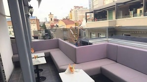 Terraza en la azotea con un área de estar moderna, que incluye un sofá y dos mesas, con vistas a un paisaje urbano.