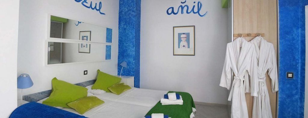 A hotel room with a bed, green pillows, a mirror, a robe rack, and a blue accent wall.