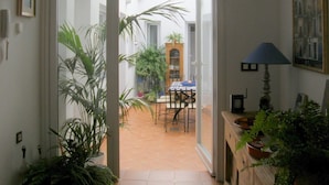 An interior view of a room with a tiled floor, a wooden cabinet with a lamp, and a door leading to a courtyard with plants and a table.
