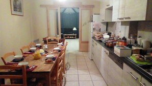 A dining area with a wooden table set for a meal, surrounded by chairs. There is a kitchen area with cabinets and appliances to the right. - Chez Louis - Chambres d'Hôtes (La Canourgue)