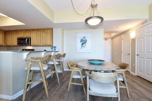 A kitchen with wooden cabinets, a microwave, a dining area with wooden chairs and a table, and a ceiling light fixture. - Spacious open-concept resort condo w/private balcony in Destin! (Destin)
