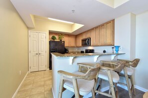 A kitchen with wooden cabinets, a black refrigerator, a microwave, and a bar area with wooden stools. - Spacious open-concept resort condo w/private balcony in Destin! (Destin)