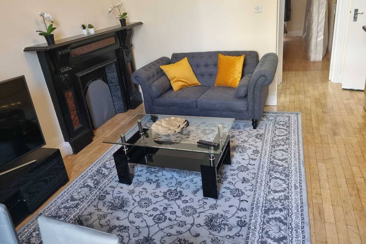 Ein Wohnzimmer mit Kamin, einem grauen Sofa mit gelben Kissen, einem Couchtisch und einem gemusterten Teppich.