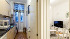 A compact kitchen with modern appliances, a dining area with a TV, and a view of the outdoors through a window with patterned curtains. - Spacious Centrally Located One Bed (London)