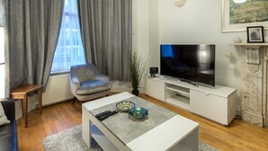 A modern living room with a flat-screen TV, grey curtains, a grey sofa, and a white coffee table with decorative bowls. - Spacious Centrally Located One Bed (London)