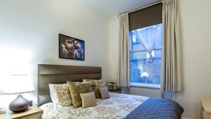 A neatly arranged bedroom with a large bed, a bedside table with a lamp, a framed picture on the wall, and a window with curtains. - Spacious Centrally Located One Bed (London)