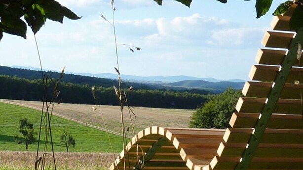 Un banc en bois, doté d’un dossier incurvé, se trouve dans un champ verdoyant, offrant une vue lointaine sur des collines.