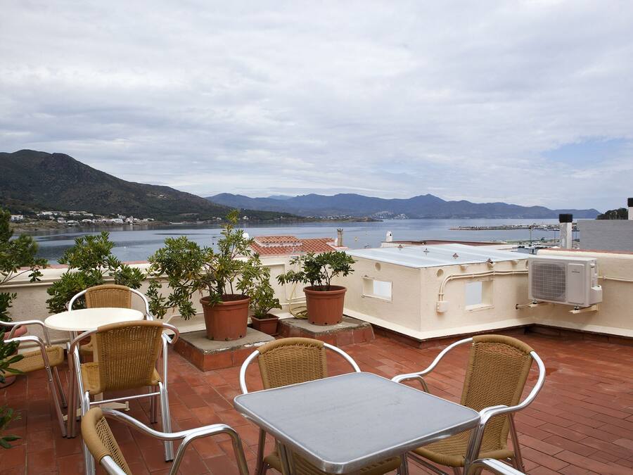 A rooftop terrace with outdoor furniture, potted plants, and a view of the sea and mountains.