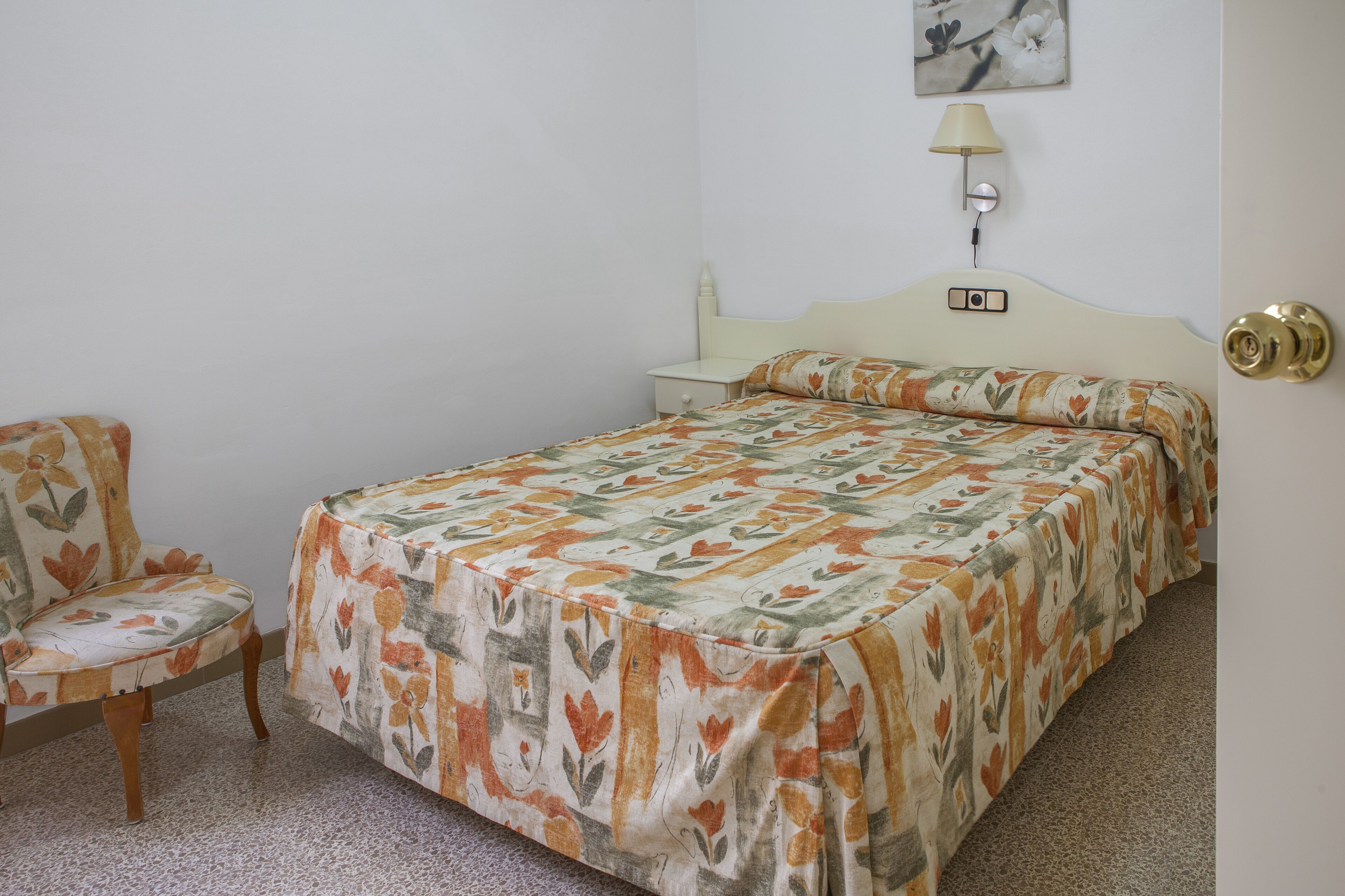 Un dormitorio con cama, una silla, una mesita de noche, una lámpara y un panel de control en la pared.