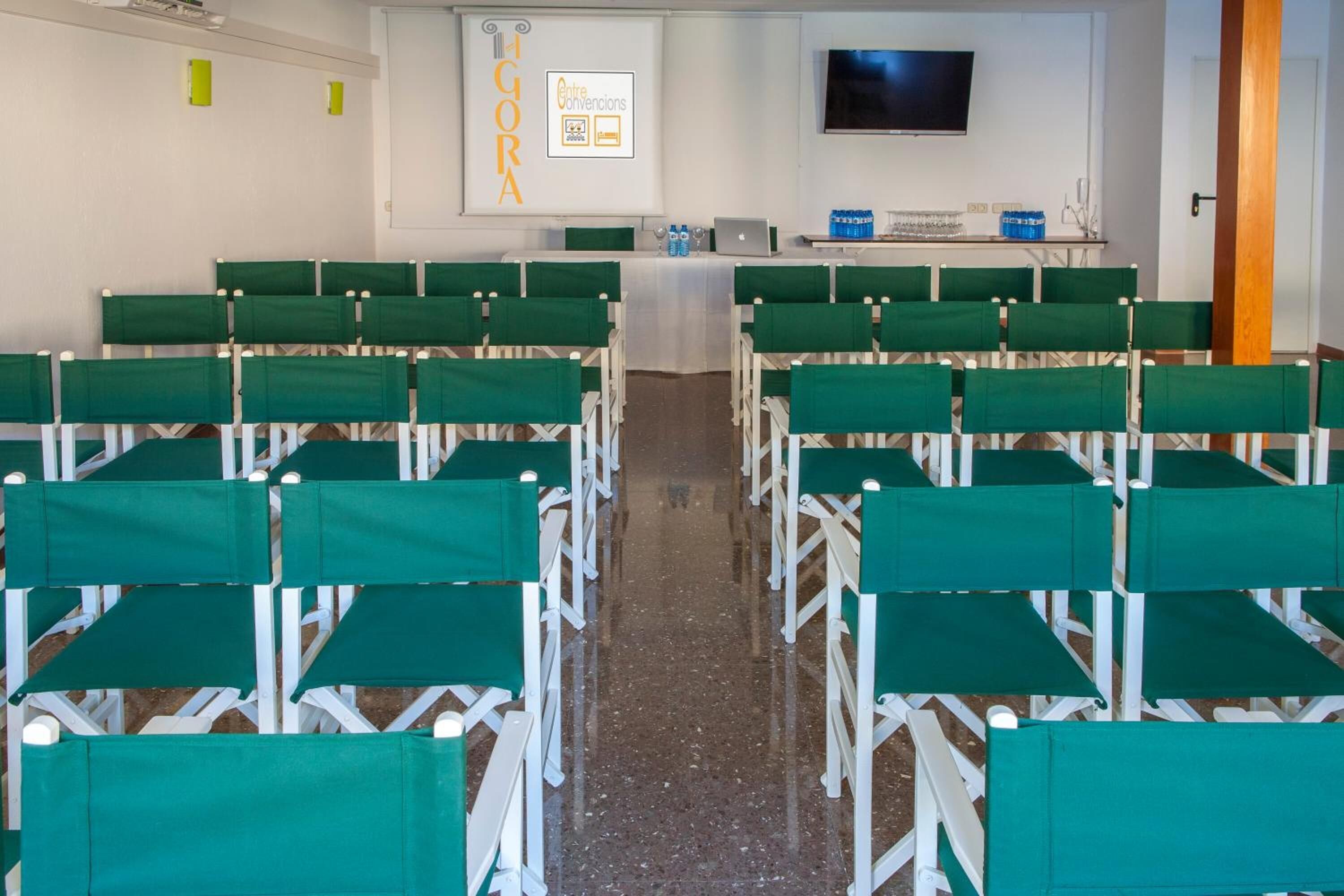 A conference room with rows of green chairs, a white table, a projector screen, and a TV on the wall.