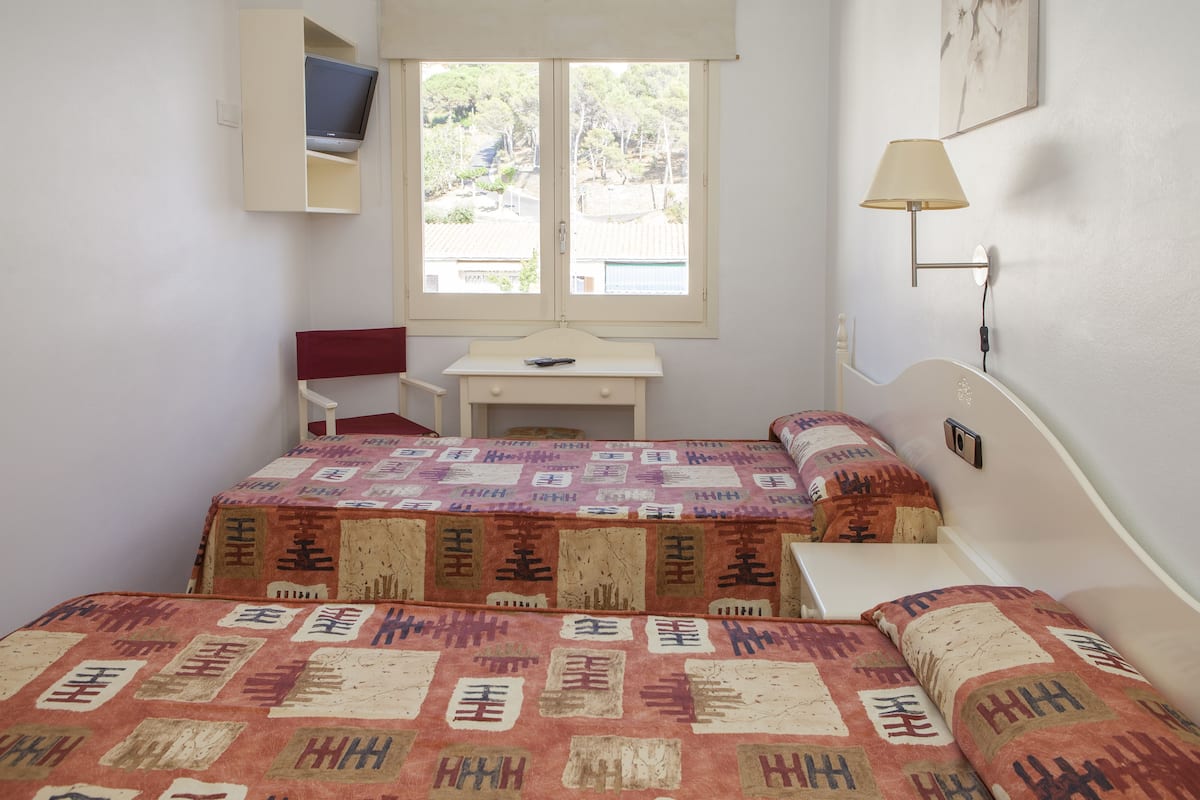 A bedroom with two beds, a desk, a chair, a window, and a wall-mounted lamp.