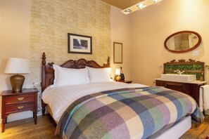 Una cama bien hecha con un edredón escocés, una mesita de noche con una lámpara, un aparador con un espejo y un baño con un lavabo verde.