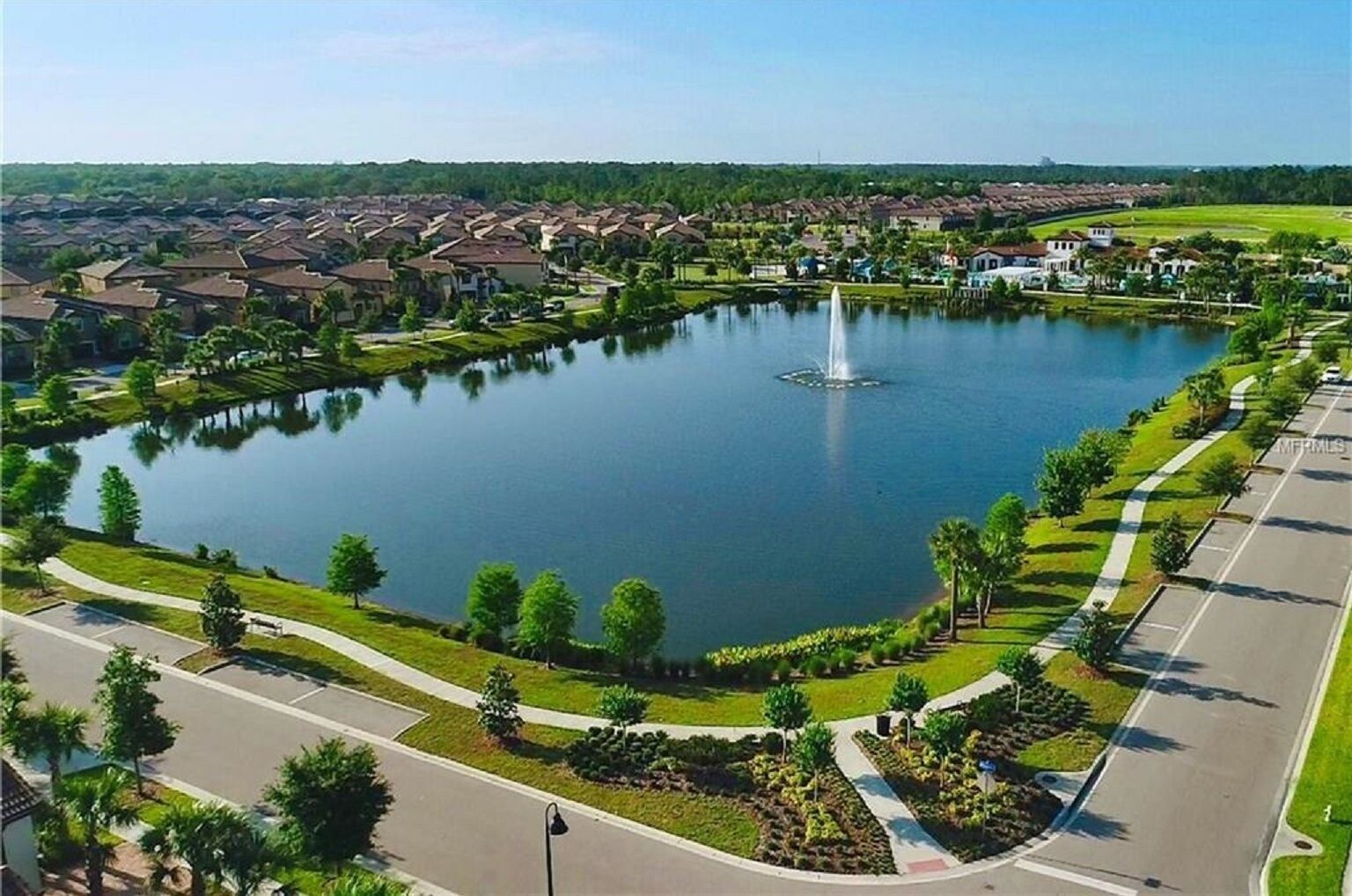 An aerial view of a residential area with a central lake, a fountain, and a well-maintained park with pathways and trees.