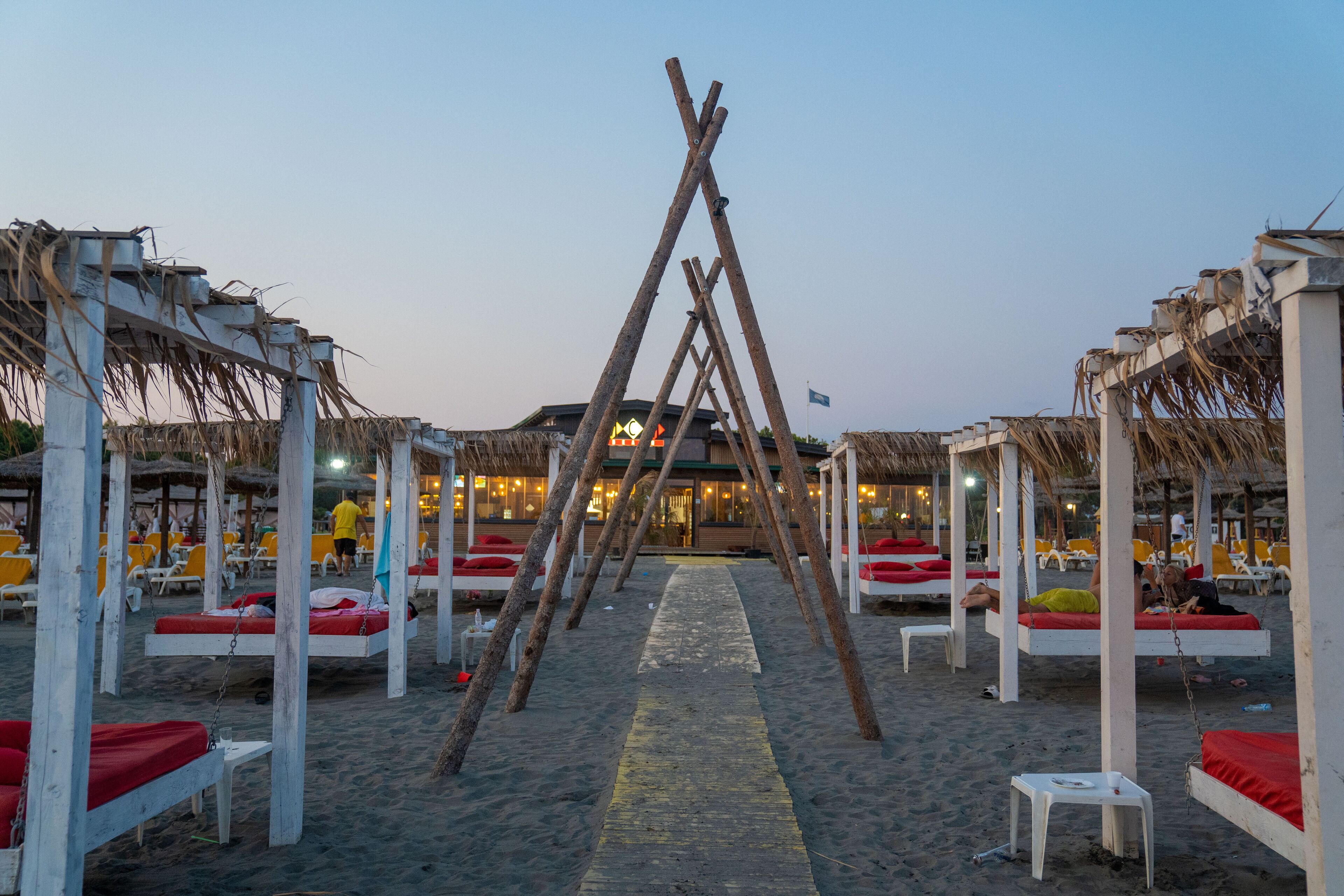 A beach with wooden structures and red cushions, a pathway leading to a building, and a flag in the distance.