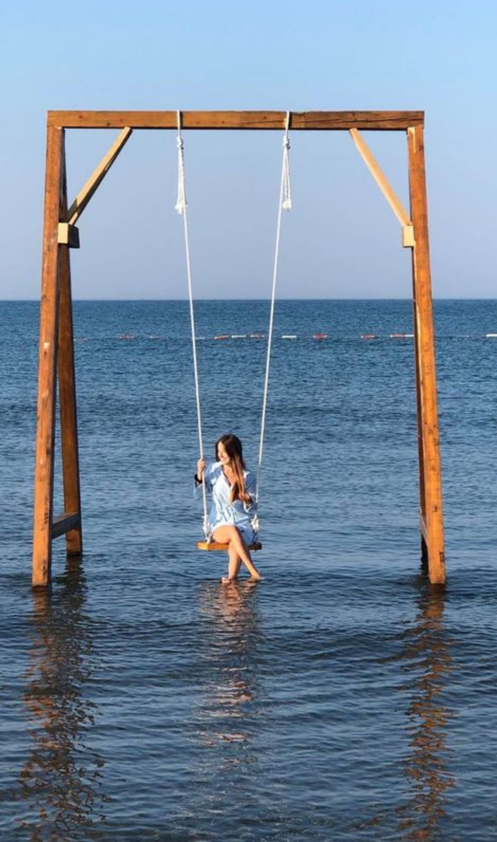 A wooden swing set over water with a clear view of the sea and boats in the distance.