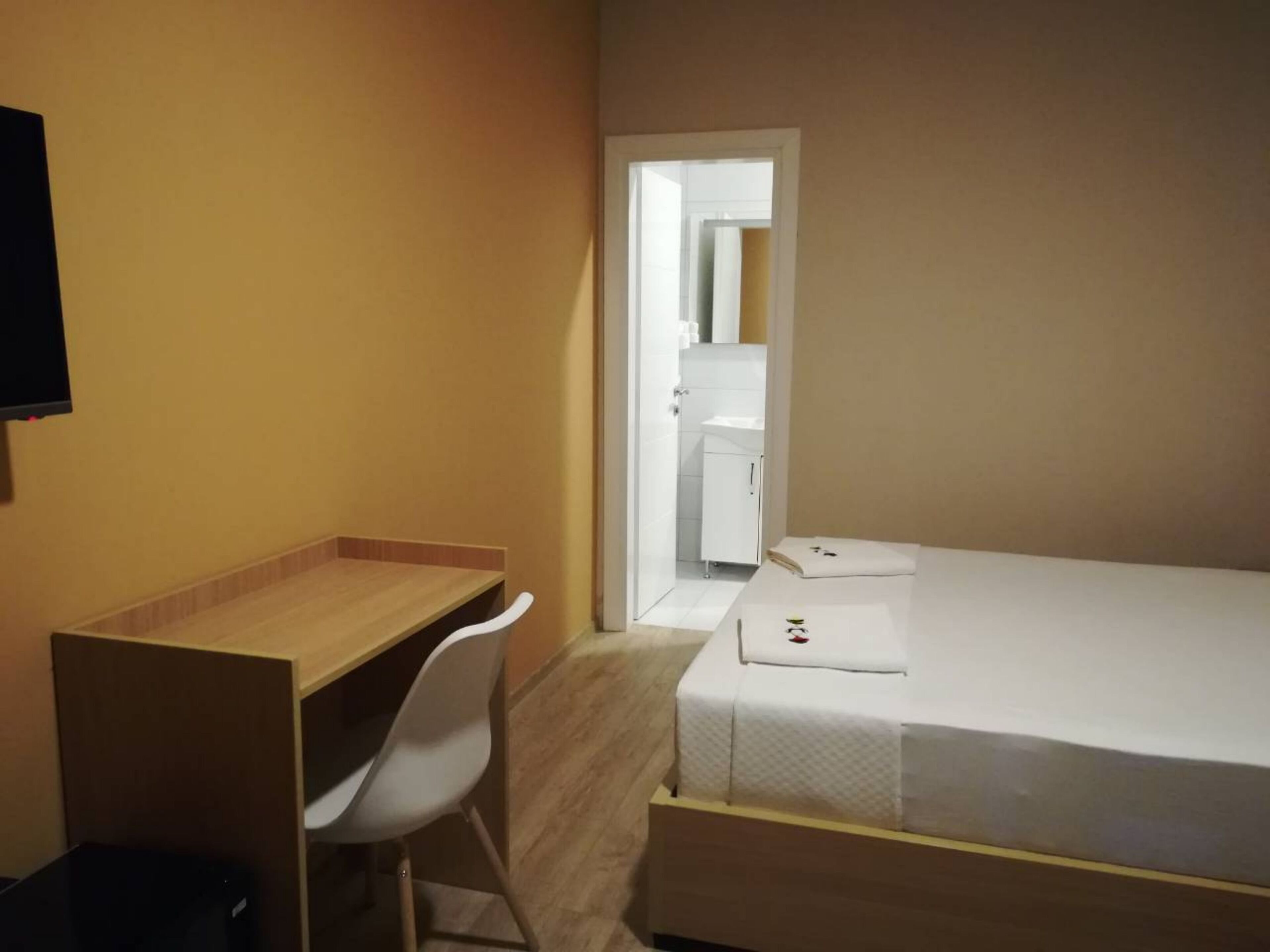 A small, single-room hotel room with a wooden desk, white chair, and a neatly made bed.