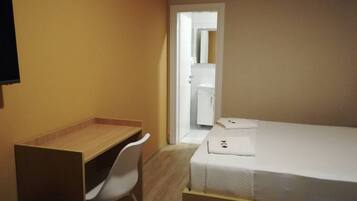 A small, single-room hotel room with a wooden desk, white chair, and a neatly made bed.