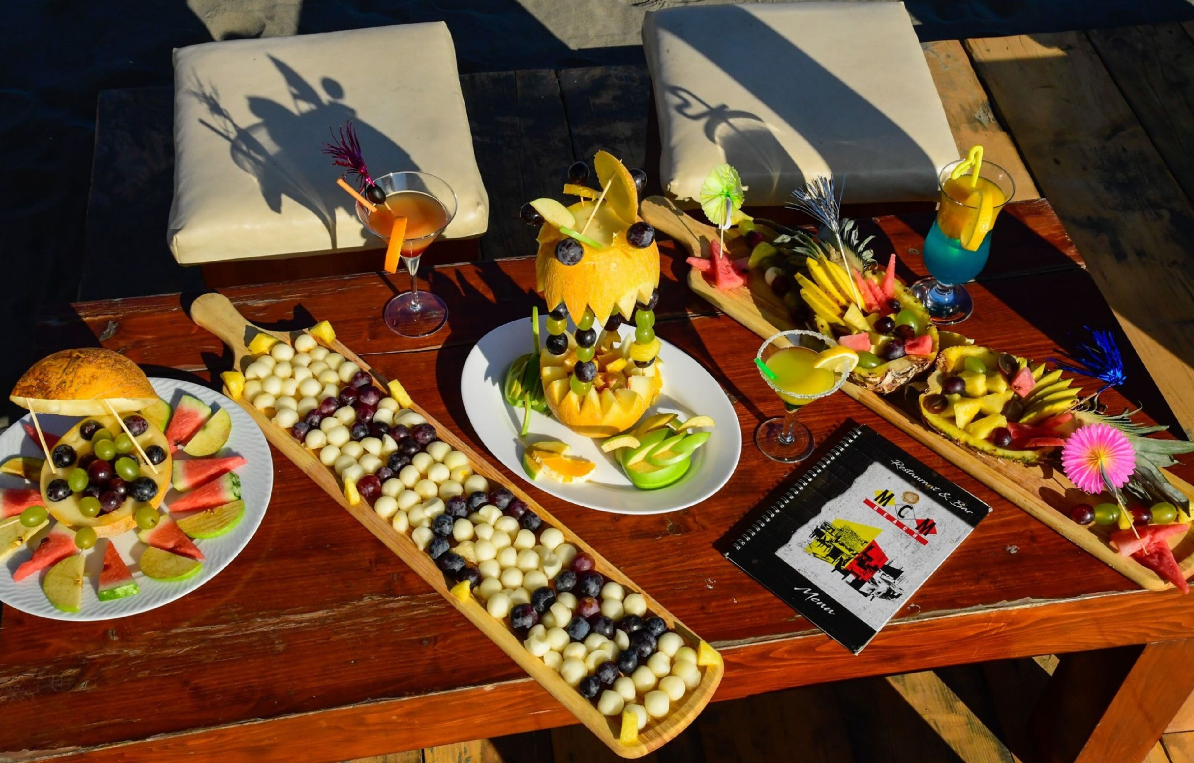 A wooden table with various fruit platters and drinks, a menu, and a notebook.