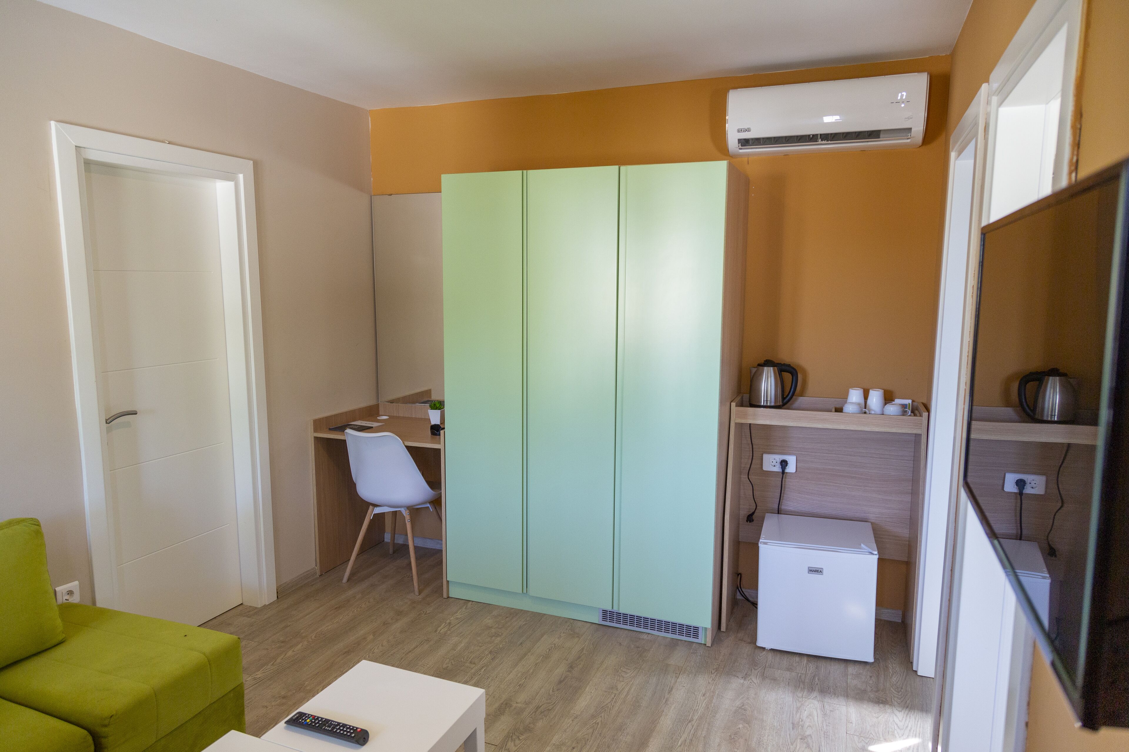 Ein kleines Zimmer mit Küchenzeile, einem grünen Sessel, einem weißen Tisch und einem weißen Kleiderschrank.
