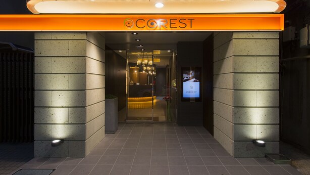 Ein Hotel Eingang mit einem gut sichtbaren 'COREST' Schild über der Tür, ein einladender Empfangsbereich ist im Inneren sichtbar, und ein modernes Design mit gefliestem Boden und einer Glastür.