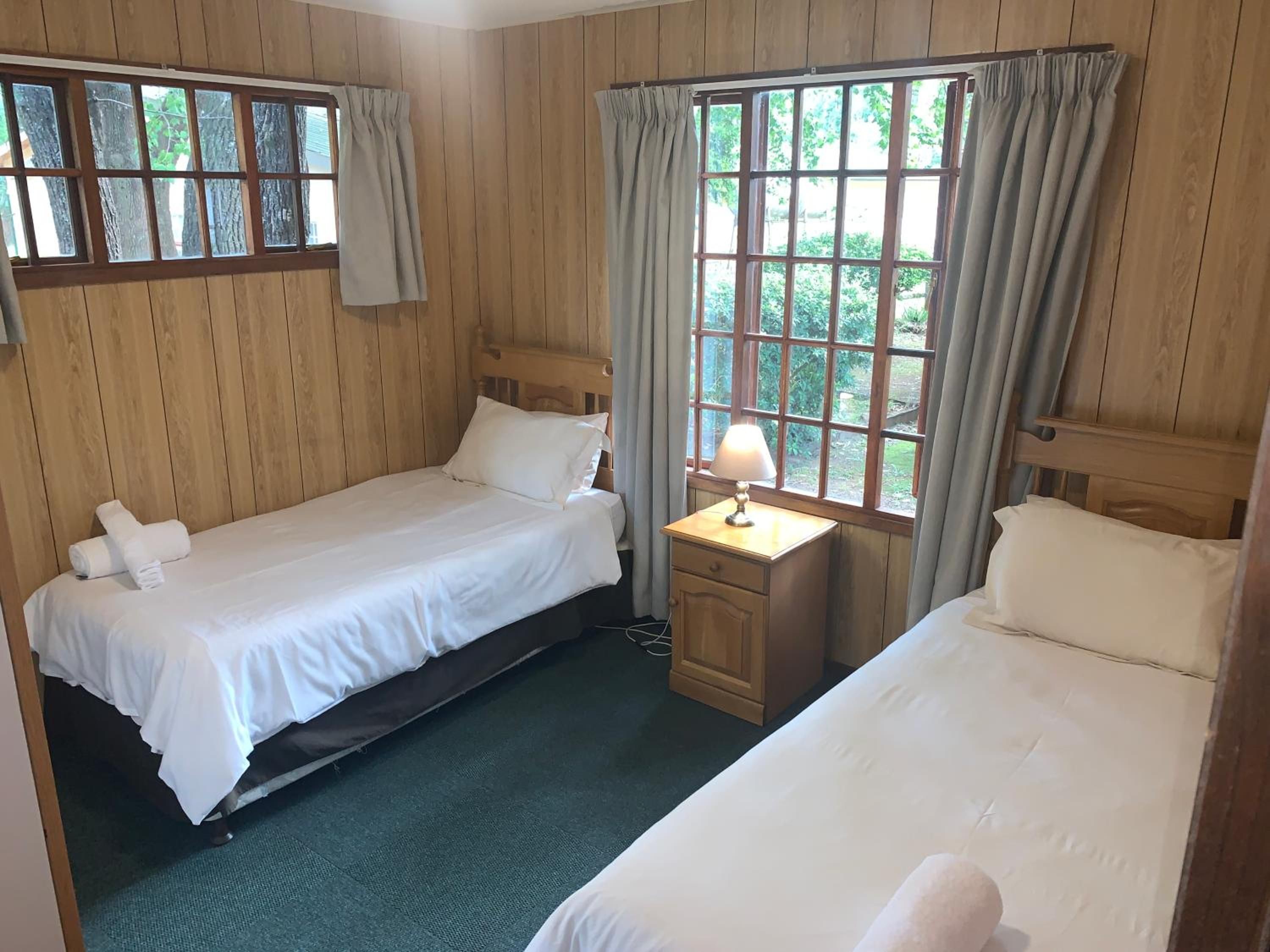A room with two beds, a nightstand with a lamp, and large windows with curtains.